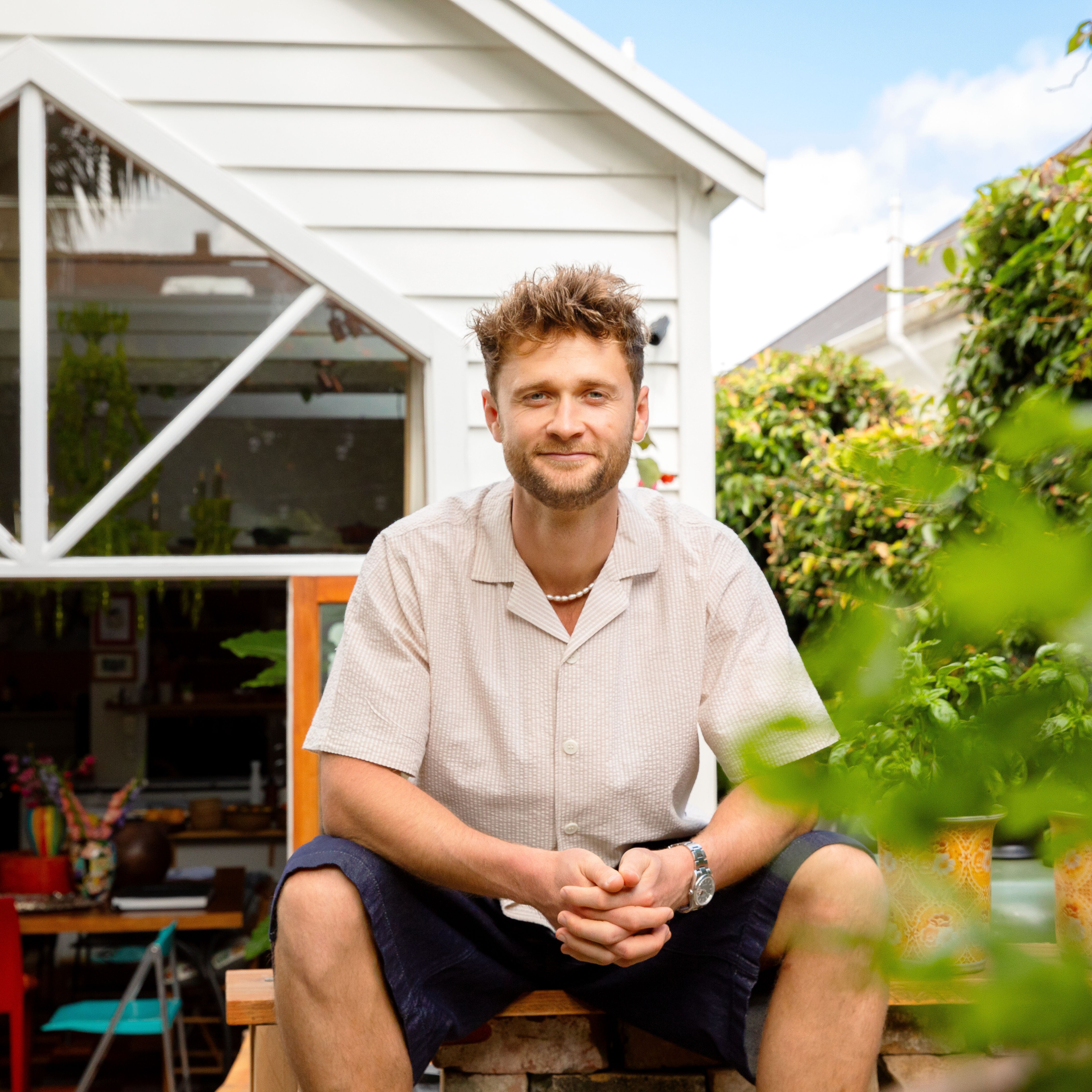 A portrait image of Hercules Noble sitting on a wooden table in his backyard, There is greenery in the foreground and right of the image. 