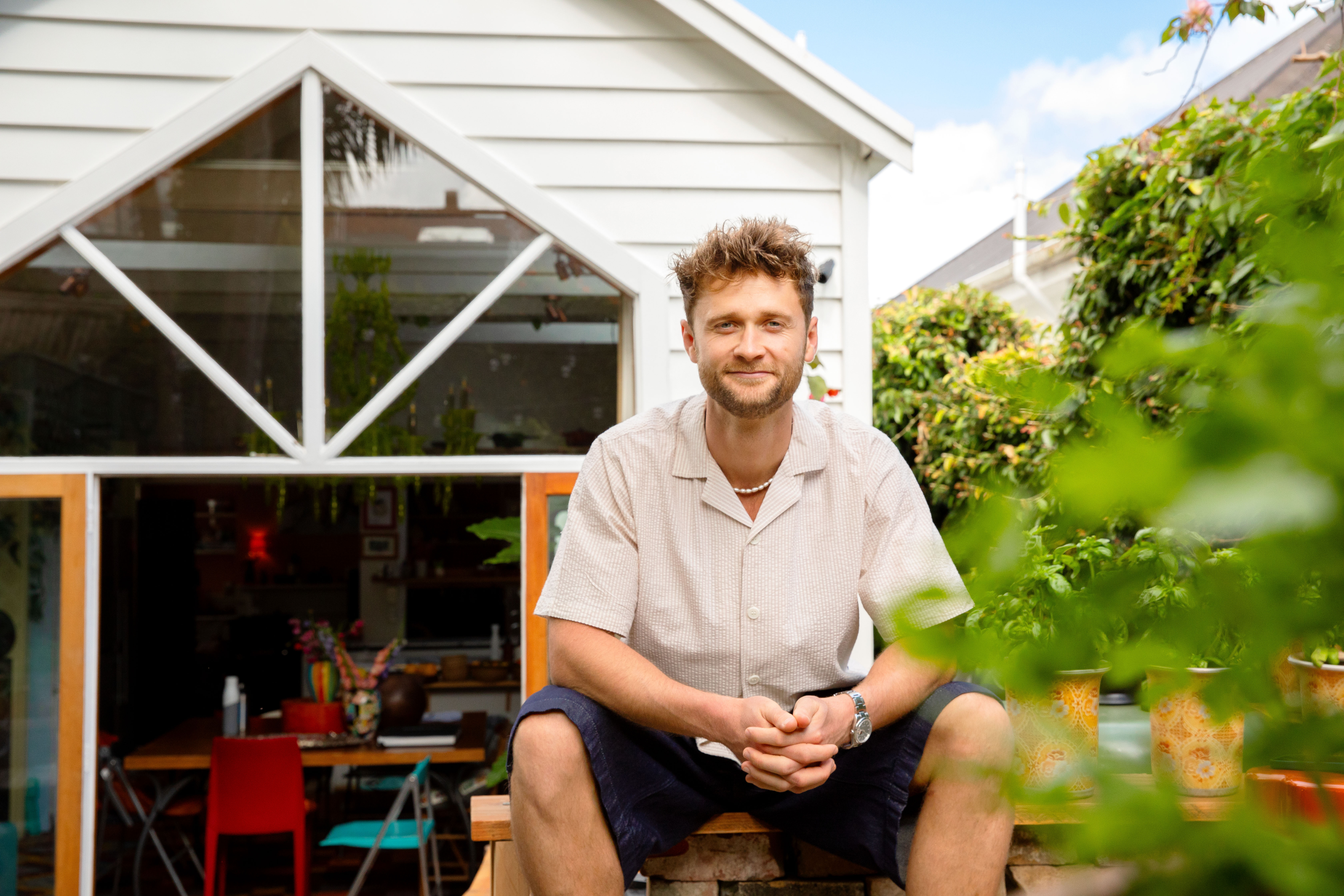 A portrait image of Hercules Noble sitting on a wooden table in his backyard, There is greenery in the foreground and right of the image. A portrait image of Hercules Noble sitting on a wooden table in his backyard, There is greenery in the foreground and right of the image.