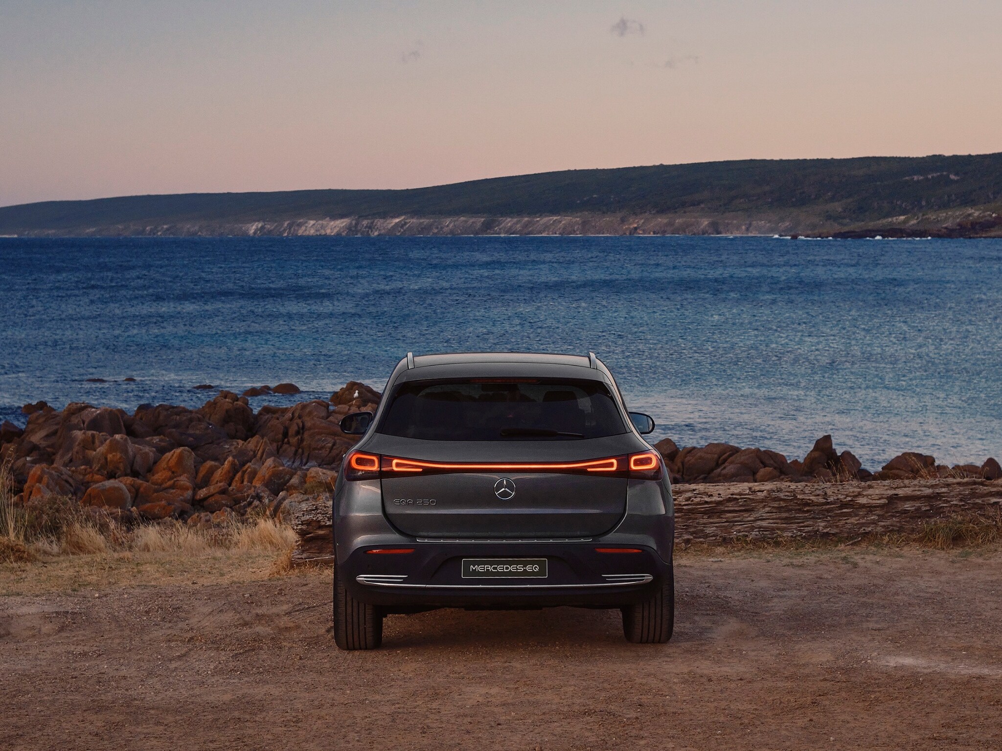 A grey EQA 250 parked in front of the ocean at dusk, pictured from behind. Image credit/photographer: Simon Shiff