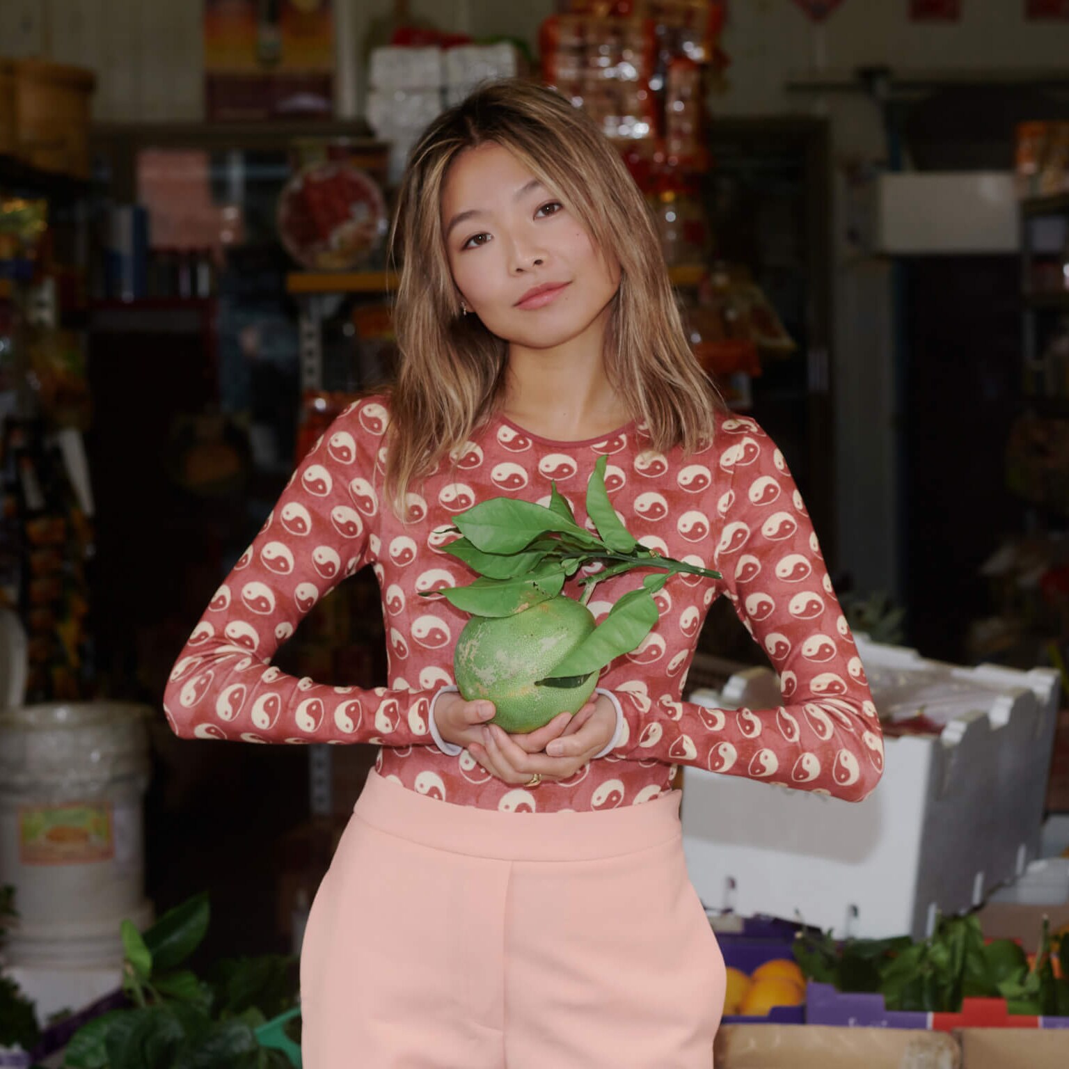 Yan Yan Chan wears a red outfit and holds fruit at an Asian grocery store.