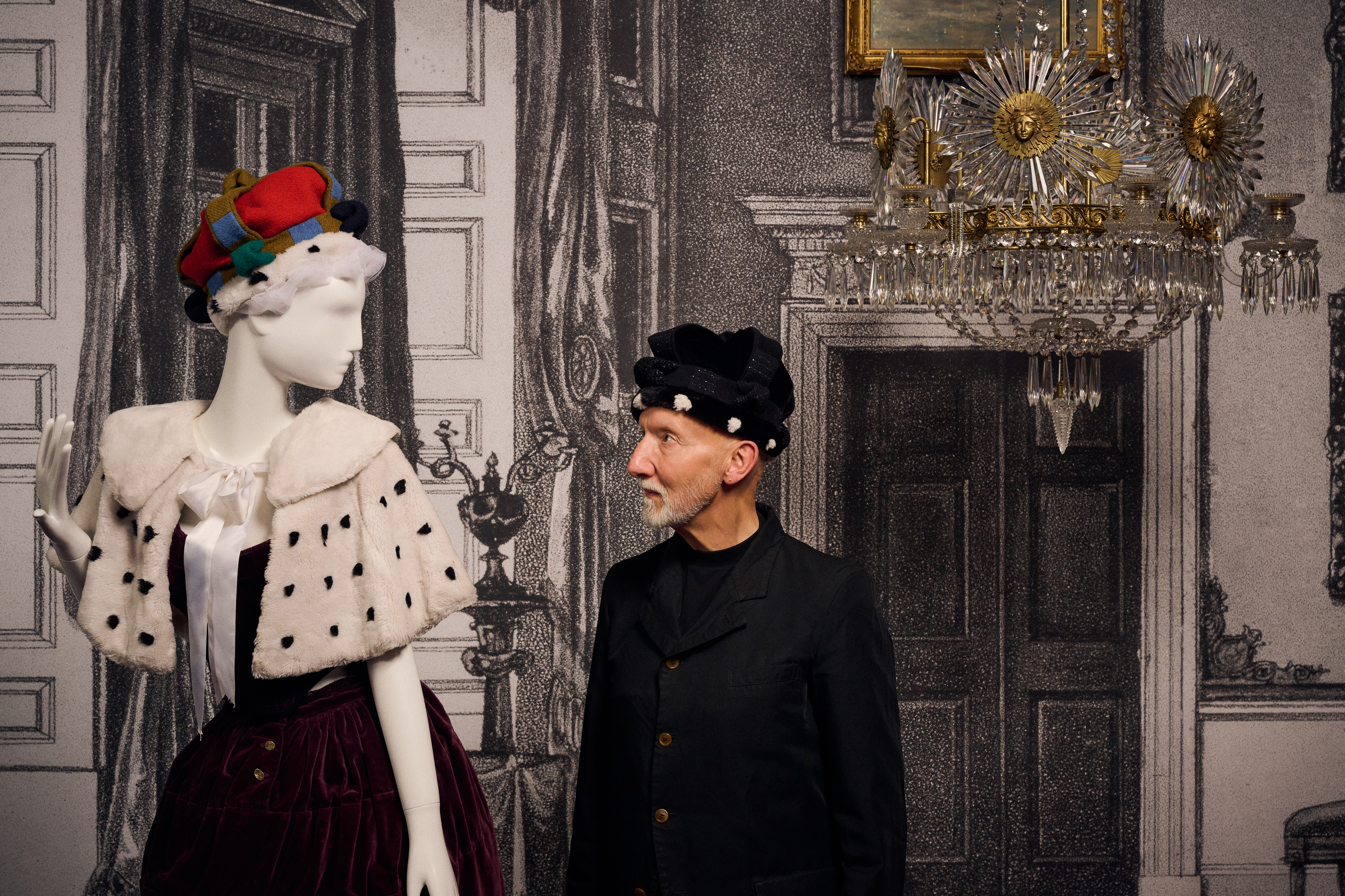 Portrait of Stephen Jones, British Milliner, with Vivienne Westwood, Ensemble, 1987, Harris Tweed collection, autumn-winter 1987–88, in Westwood | Kawakubo on display from 7 December 2025 to 19 April 2026, at NGV International, Melbourne. Photo: Eugene Hyland A bearded man in a hat, milliner Stephen Jones, looks at one of his designs for Vivienne Westwood.