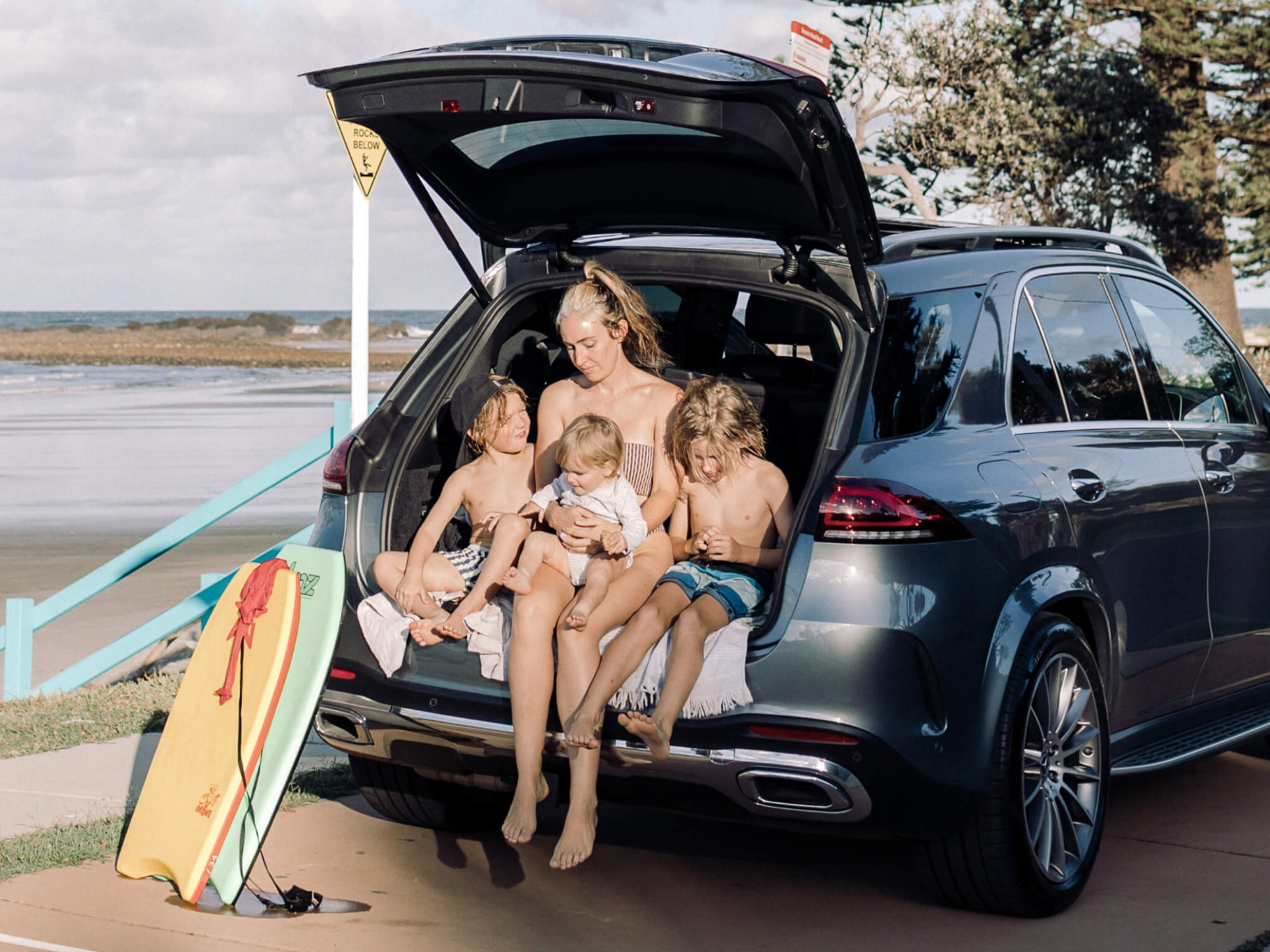 Mother and three children sitting in the boot of a Mercedes-Benz GLE SUV with boogie boards at the beach. Image credit/photographer: Tim Coulson