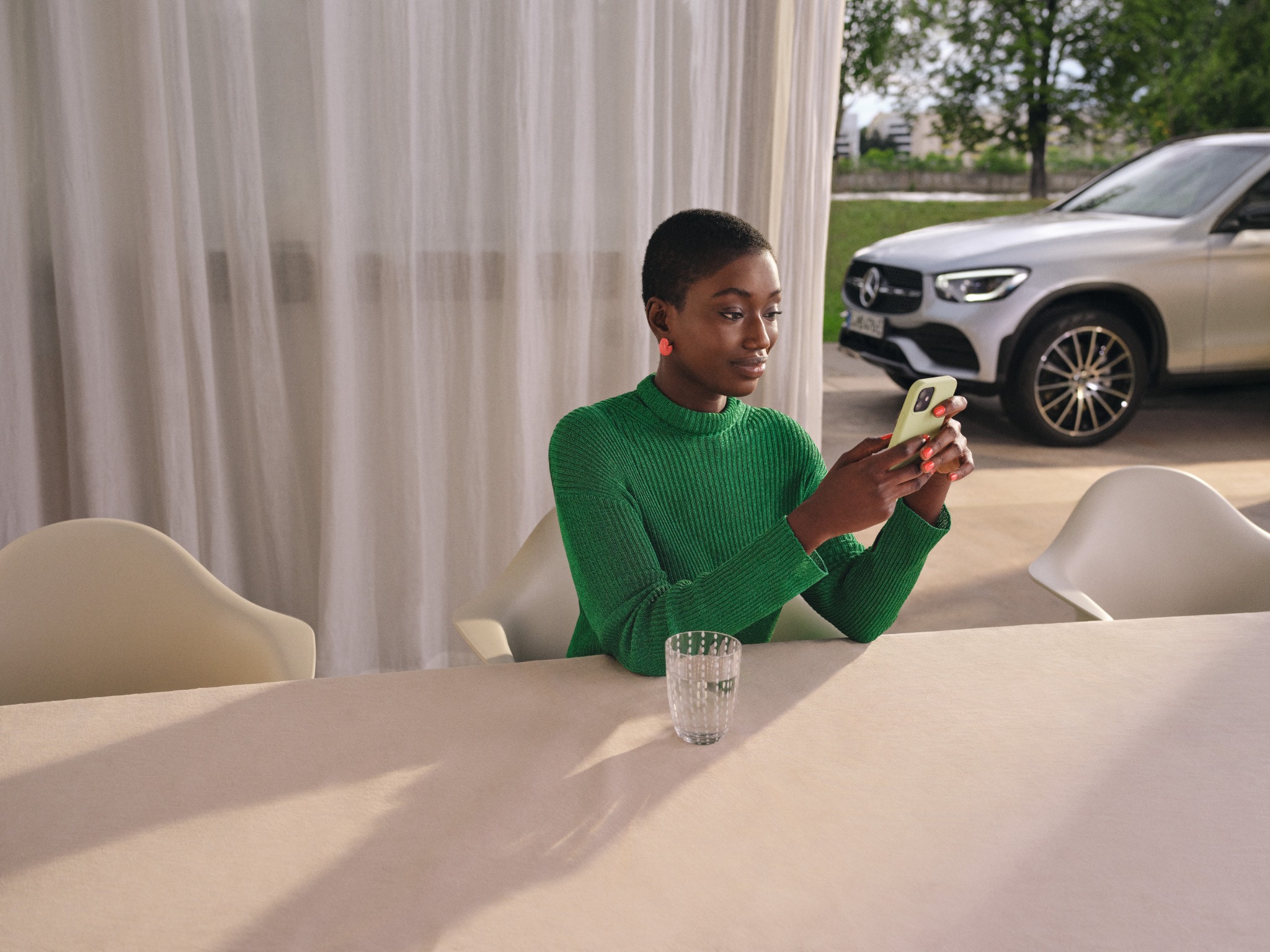A woman sitting at a table on her phone with a Mercedes-Benz vehicle in the background. A woman sitting at a table on her phone with a Mercedes-Benz vehicle in the background.