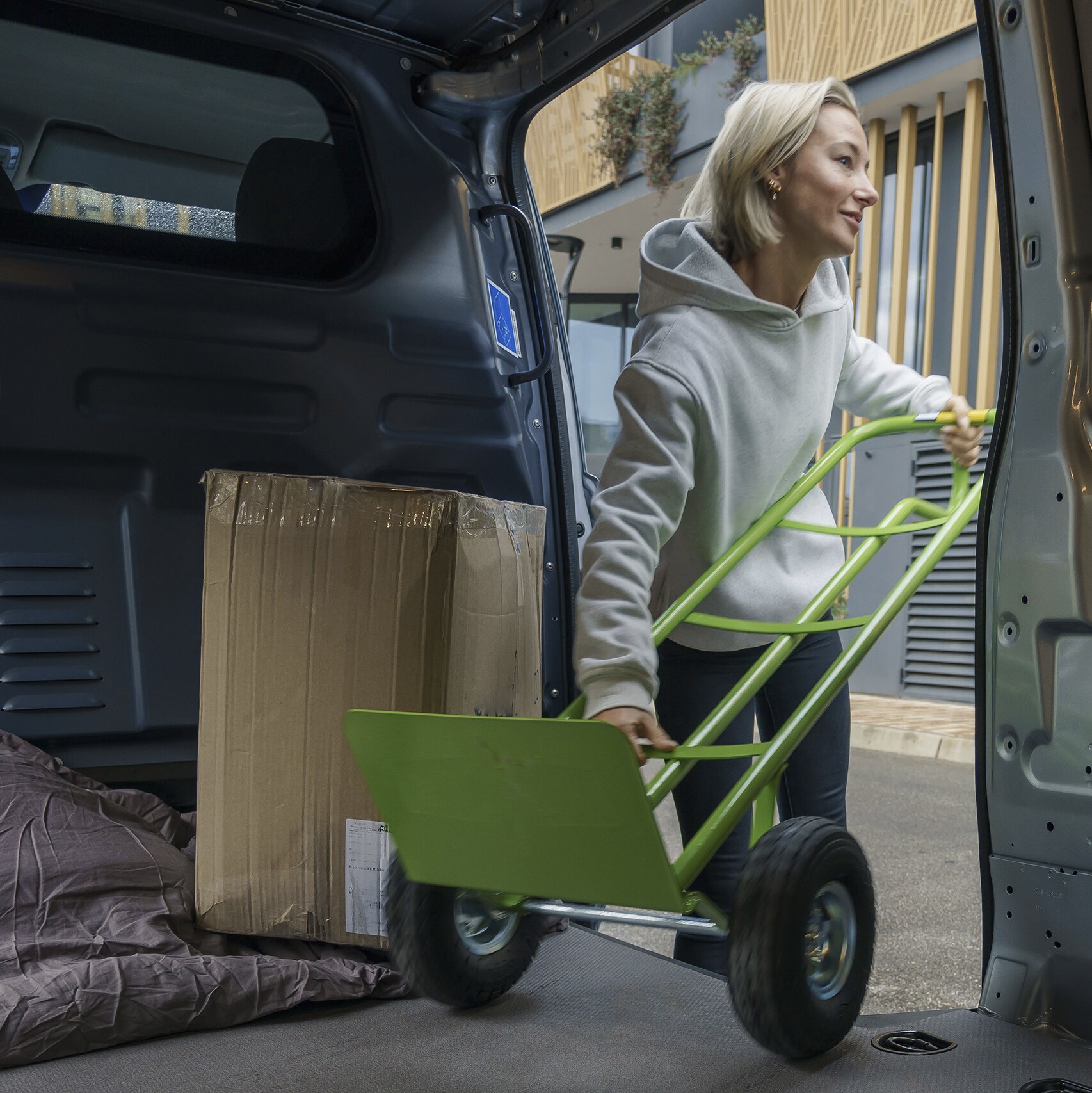 A woman in a grey hoodie loads a cardboard box into the back of a van using a green hand trolley, where a blanket covers the floor.