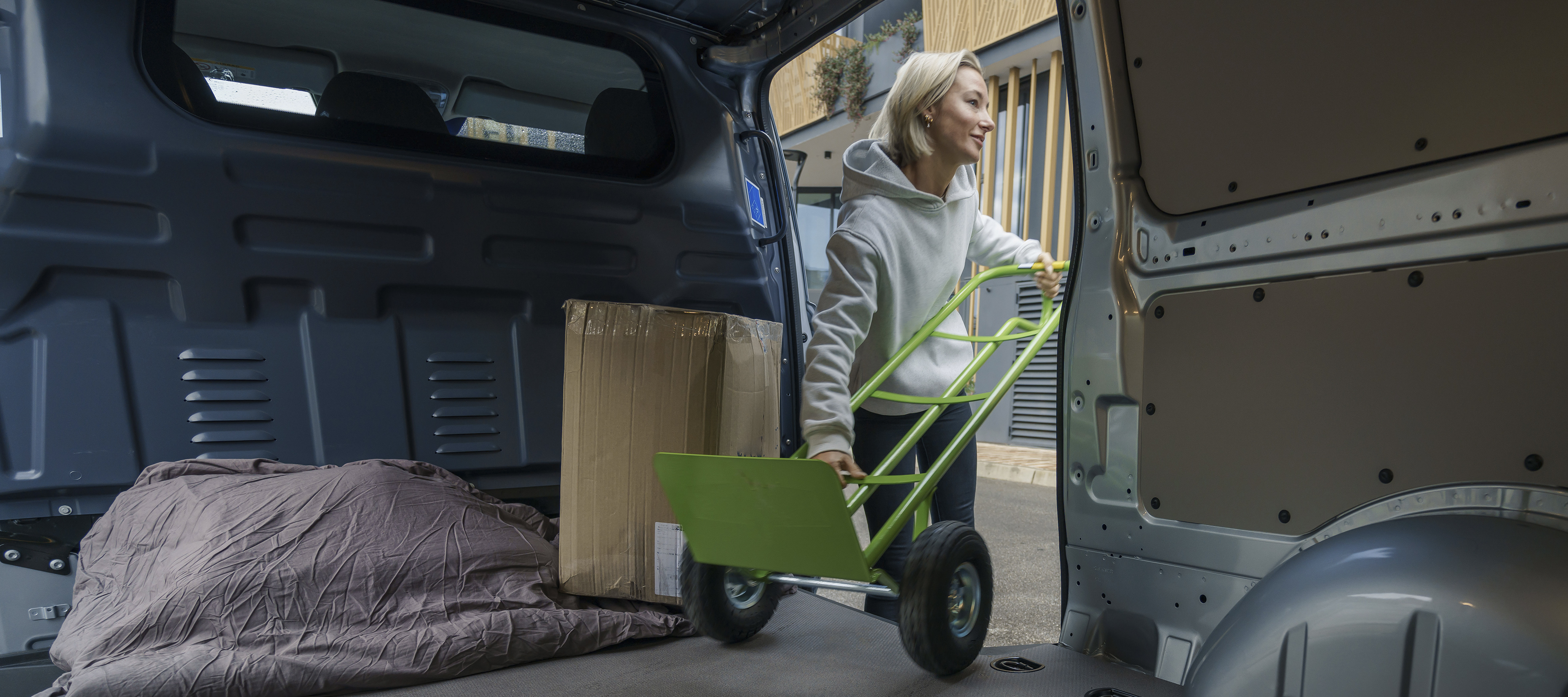 A woman in a grey hoodie loads a cardboard box into the back of a van using a green hand trolley, where a blanket covers the floor. A woman in a grey hoodie loads a cardboard box into the back of a van using a green hand trolley, where a blanket covers the floor.