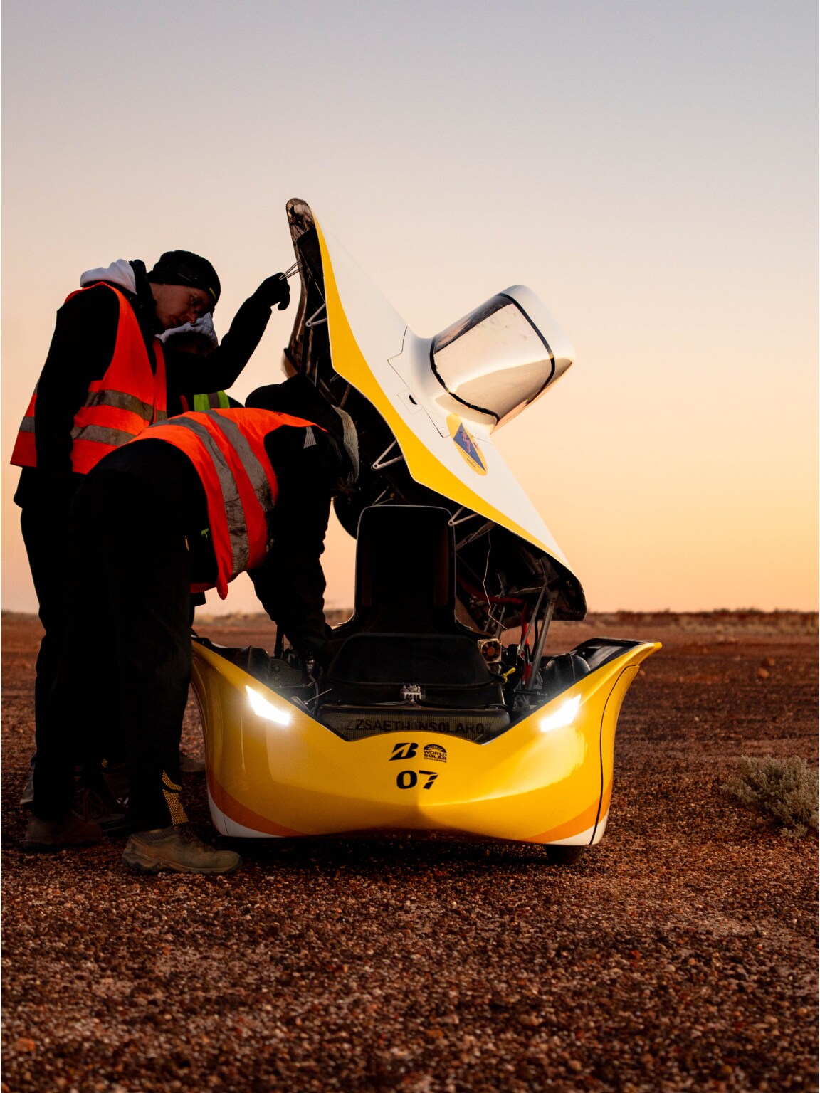 Yellow-and-white solar race car beside a black Mercedes-Benz V-Class on a desert road.