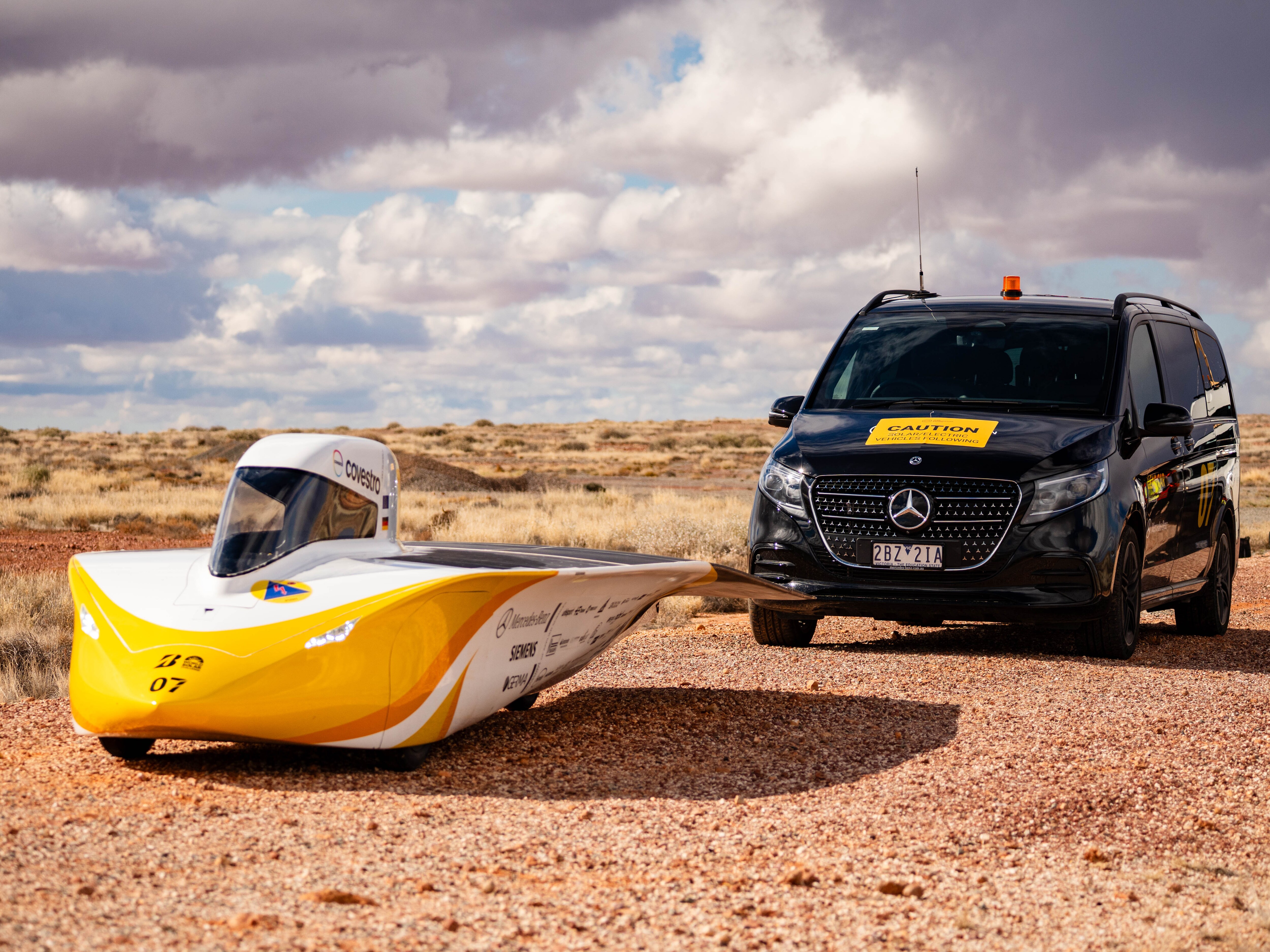 Yellow-and-white solar race car beside a black Mercedes-Benz V-Class on a desert road.