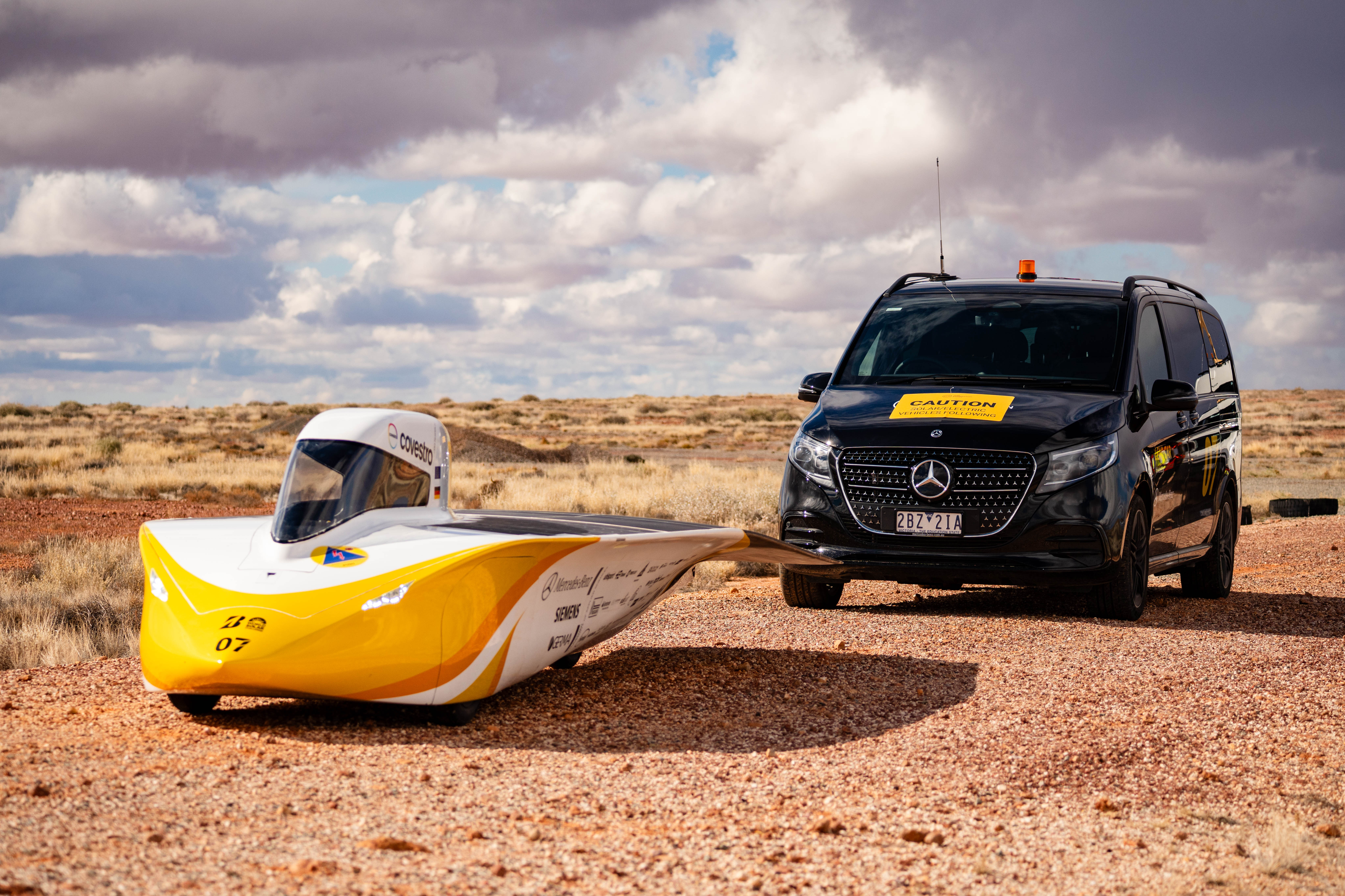 Team Sonnenwagen Yellow-and-white solar race car beside a black Mercedes-Benz V-Class on a desert road.