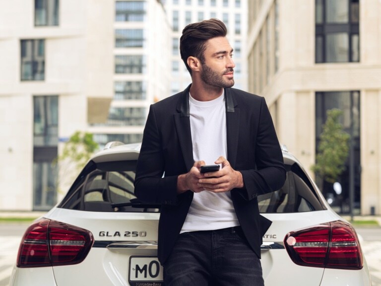 A man leans against his Mercedes-Benz GLA and uses the Mercedes-Benz App on his smartphone and the Navigation Digital Extras to find out what the weather will be like at his destination.